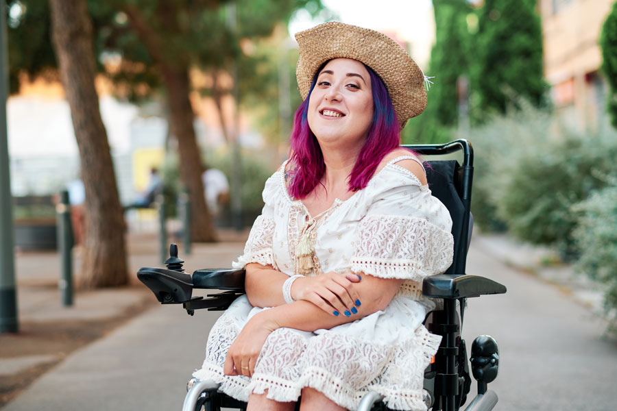 Femme souriante aux cheveux violets et en robe en dentelle assise dans un fauteuil roulant sur un trottoir ensoleillé.