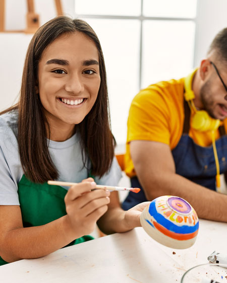 A smiling girl wearing a greet apron and boy in a bright yellow shirt and blue apron painting pottery