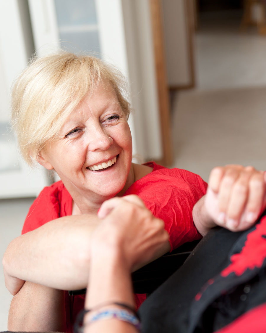 Une femme agée blonde, en chemise rouge, souriant et jouant avec quelqu’un