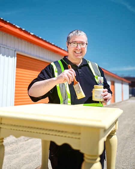 A smiling man holding a paint brush and jar of yellow paint while painting an old table yellow.