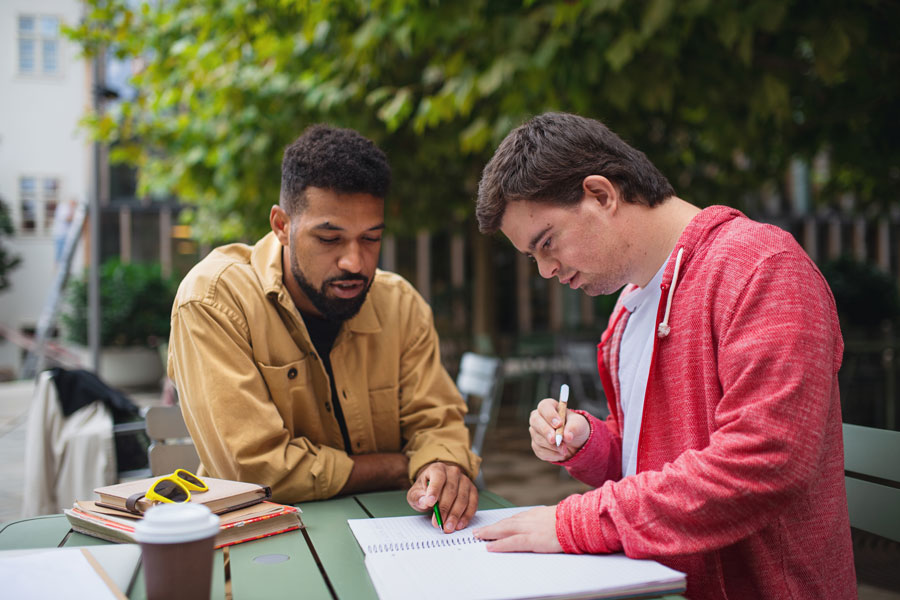 Two men at a picnic table. One is helping the other who is writing in a notebook.