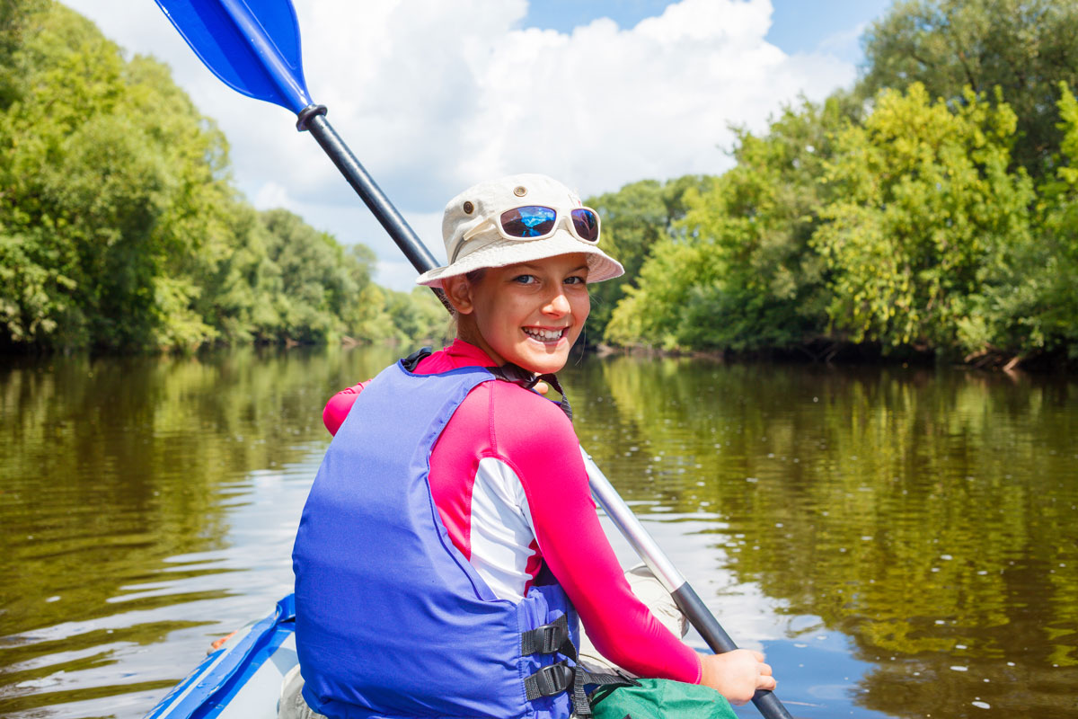 A young smiling girl wearing a bright pink top under a lifejacket while rowing a Kayak. She is wearing a bright pink top under a lifejacket