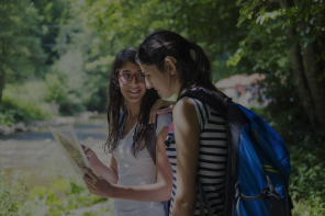 Two teen girls wearing backpacks outside reading paper directions.