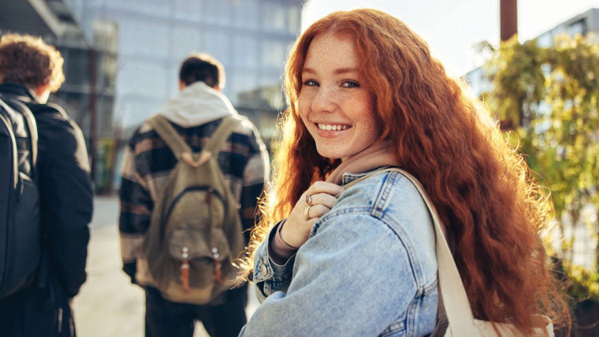 A young adult woman with red hair wearing a jean jacket and carrying a bag with a large glass building in the background. Two young men walk in front of her.