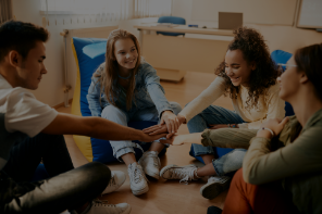 Four young people sitting in a circle of the floor putting their arms together in the middle.