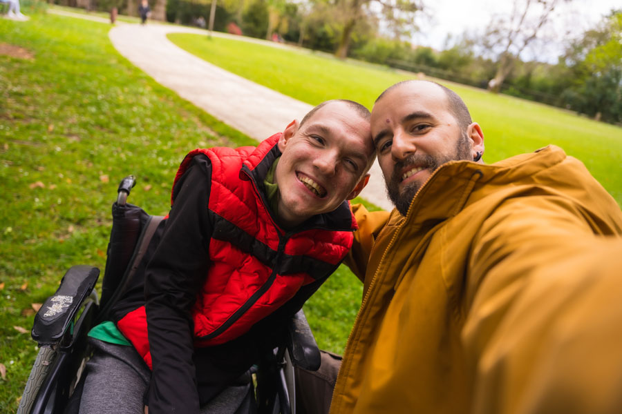 Two men at a park, one in a wheelchair, smiling for the camera.