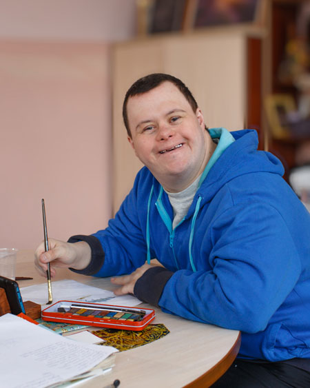 Un homme souriant, portant un pull-over bleu, assis à une table avec des fournitures de peinture et de dessin.