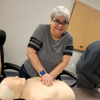 A woman with grey hair practicing CPR on a half mannequin's chest.