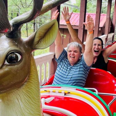Two woman riding a bright red roller coaster with a deer head at the front. They are smiling and have their arms in the air.