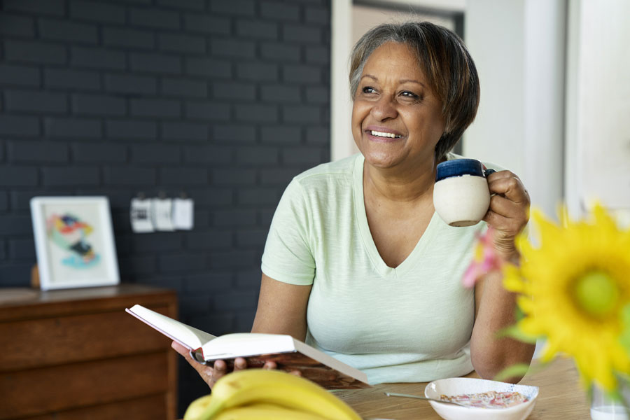 A brown skinned woman holding a coffee mug and looking at a book while sitting at a table.