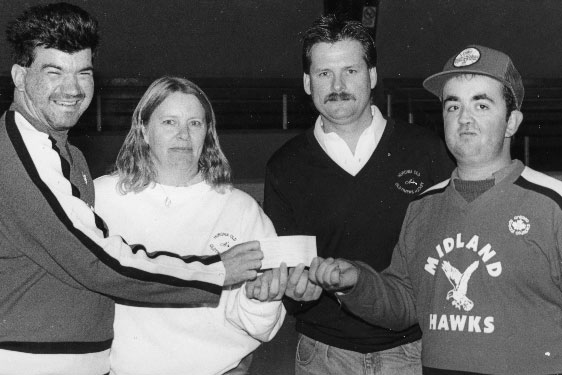 A man presenting a cheque to another man with a woman and man in the background inside the Midland arena