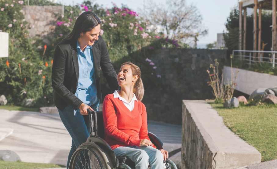 Jeune femme souriante en chandail rouge, dans un fauteuil roulant, poussée par une autre femme.