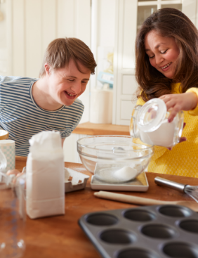 A woman is pouring milk into a bowl of flour while a younger adult smiling man watches. .
